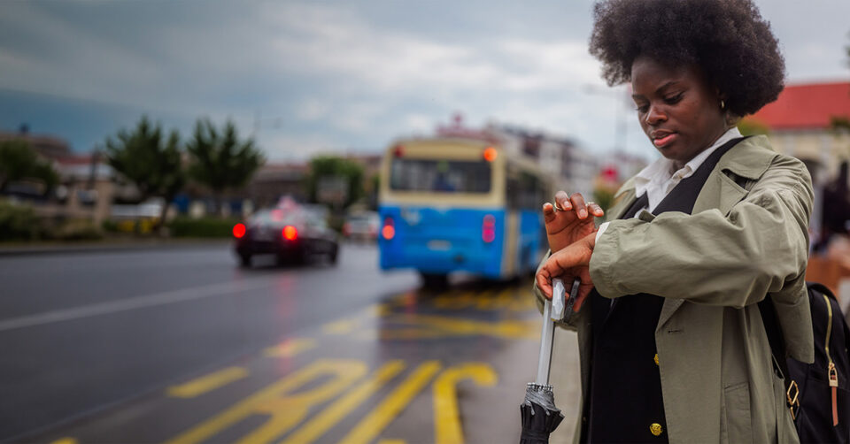 Woman checks her watch on a rainy city street as a blue bus passes in the background. 0:10 scene with traffic nearby and a backpack on her shoulder, ready to board or wait for transit.